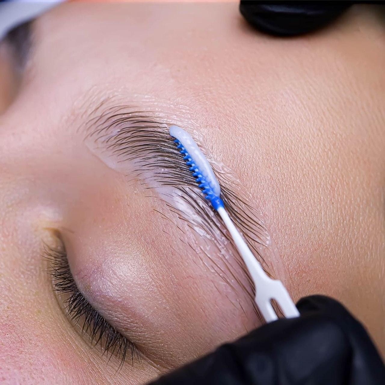 A beautician applies paint to a kleintki's eyebrows after laminating and adjusting her eyebrows